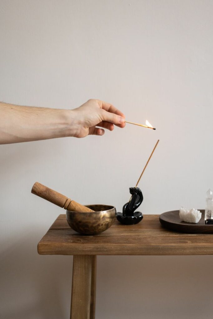 A hand lighting an incense stick with a matchstick on a minimalist wooden table with a singing bowl.